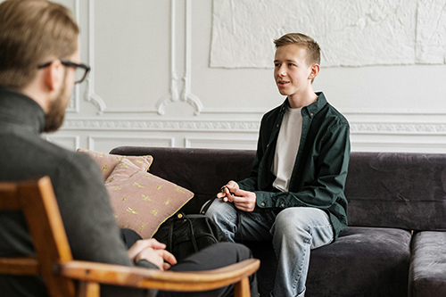 A man sitting in a chair with glasses talking to a younger man on a couch