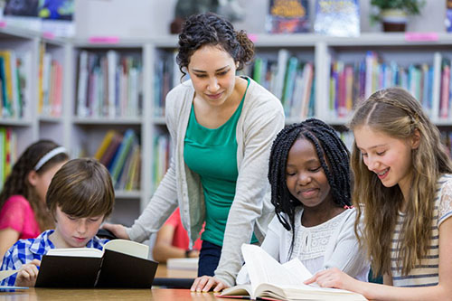 A teacher with brown hair, a green shirt and a gray sweater is teaching four students at a table