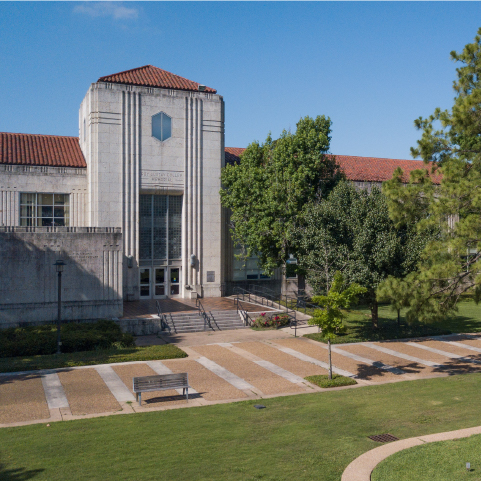 An aerial view of a wide, grey stone building with terra cotta gabled roofs against a blue sky. The building’s wide stairs lead down to a large brick walkway, grass and trees.