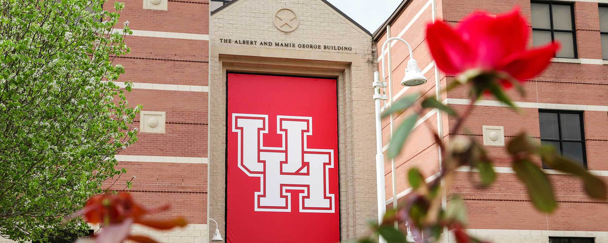 Red UH Banner haning on a brown brick building. A blurred tree branch with leaves is in the foreground