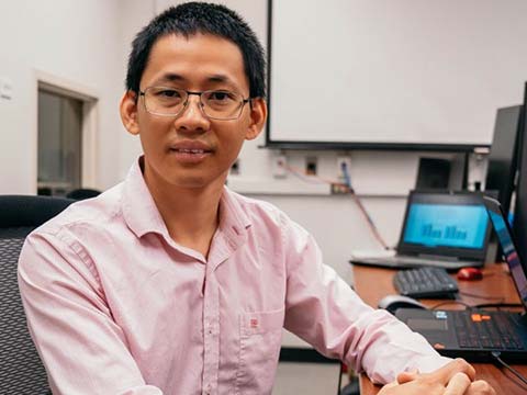 An Asian man in glasses and a pink dress shirt sits at a desk with laptops and a desktop computer.