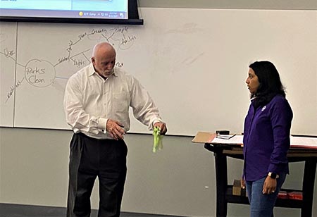 A man in a white dress shirt stands at the front of a class next to student in purple dress shirt.