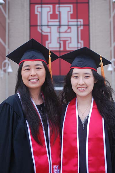 Two Asian women in black graduation regalia smile together. A large red UH banner is on the building behind them.