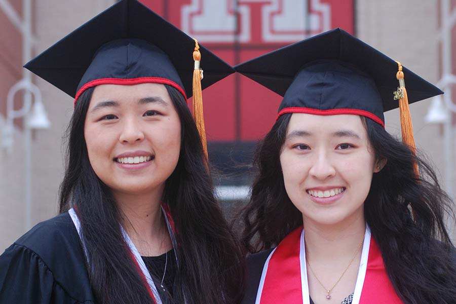 Two Asian women in black graduation regalia smile together. A large red UH banner is on the building behind them.