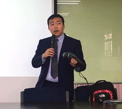 An Asian man in a suit speaks into a microphone while holding a virtual reality headset in his hand.