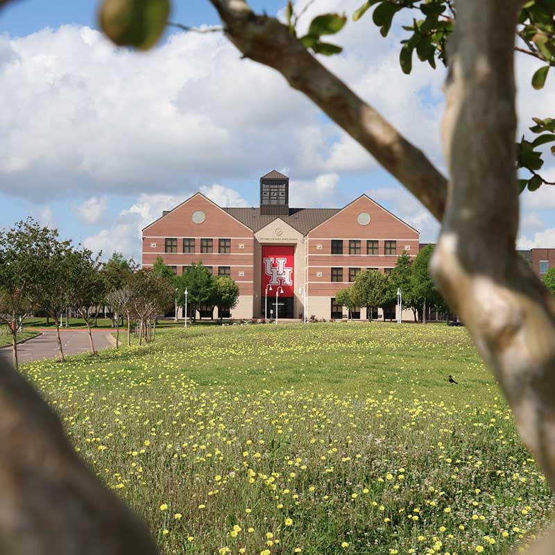 Photograph of three story brick building with a red UH banner above its front entrance. Trees surround the building and a field of flowers is in the foreground.